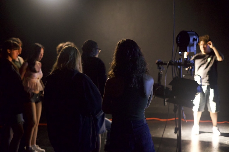 Students stand in a dark studio space while an instructor demonstrates a stage light technique